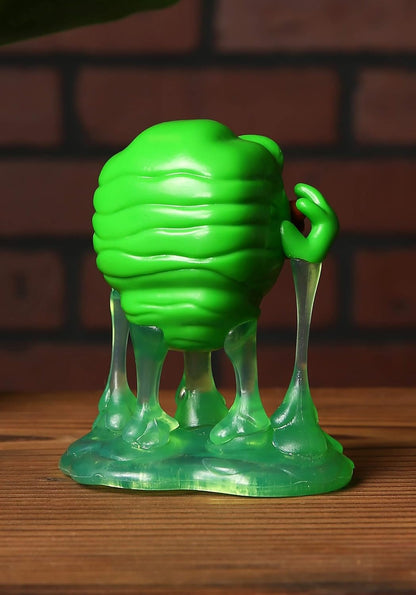 Green plastic toy figure with a textured body and transparent legs on a wooden surface with a brick wall background.