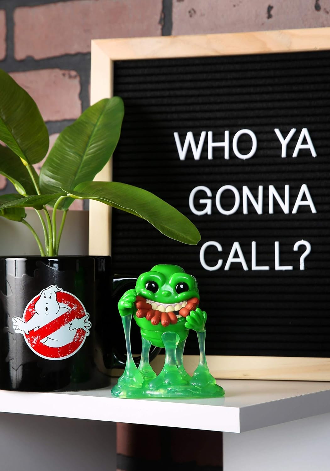 Green Slimer figurine from Ghostbusters next to a blackboard with 'WHO YA GONNA CALL?' text, plant, and Ghostbusters logo.