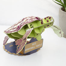 3D paper model of a hawksbill turtle on a white surface with a plant in the background