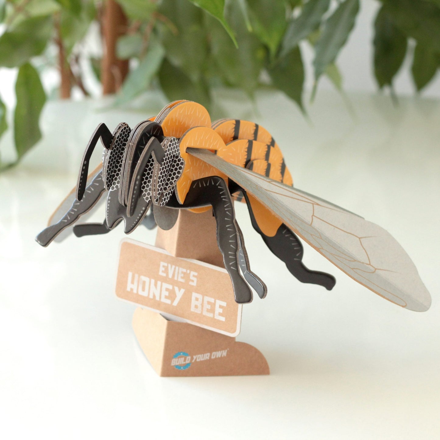 Honey bee model on a stand with 'Evie's Honey Bee' label, surrounded by green leaves.