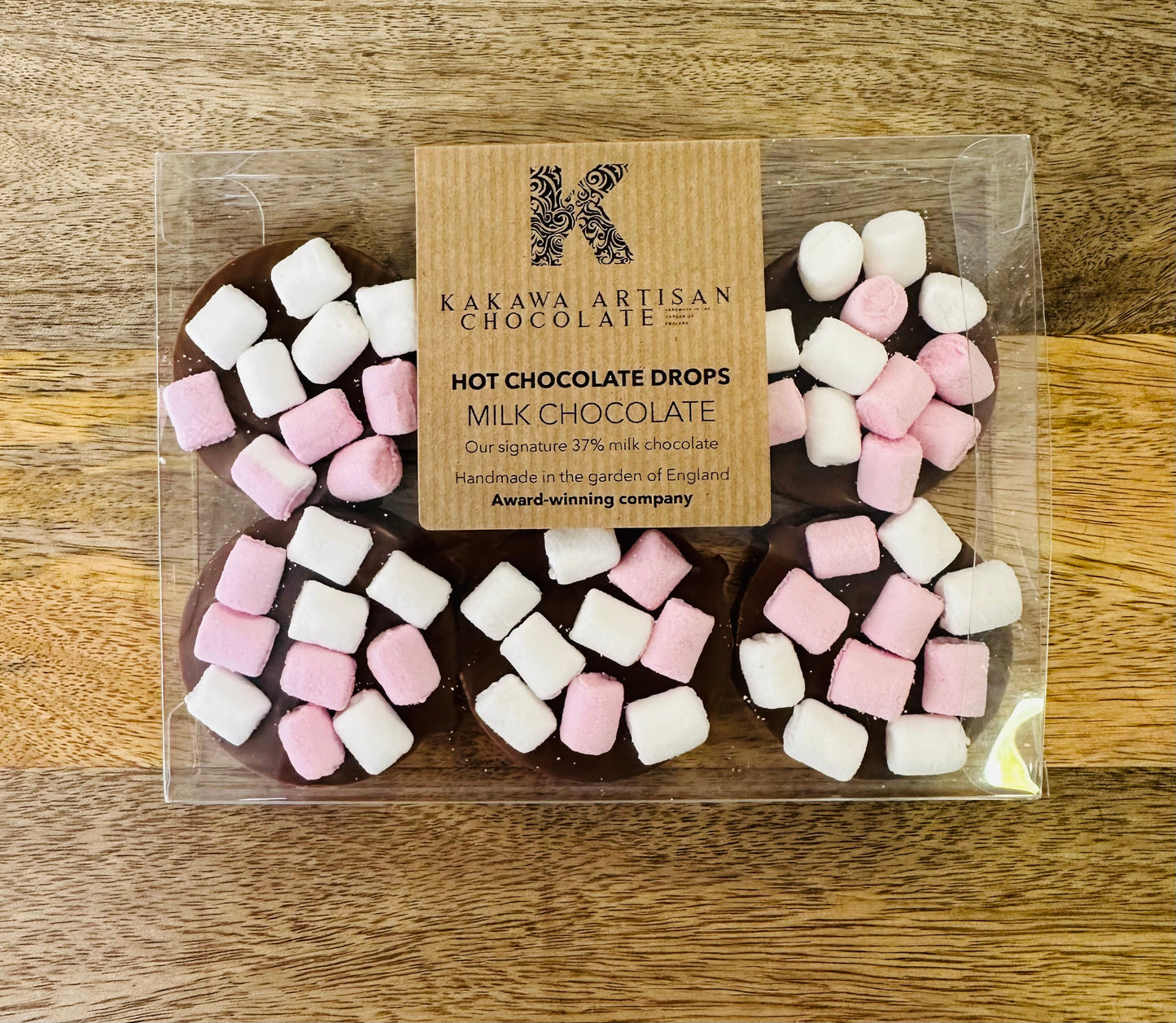 Packaged milk chocolate hot chocolate drops with marshmallows on a wooden surface, featuring Kakawa Artisan Chocolate branding.