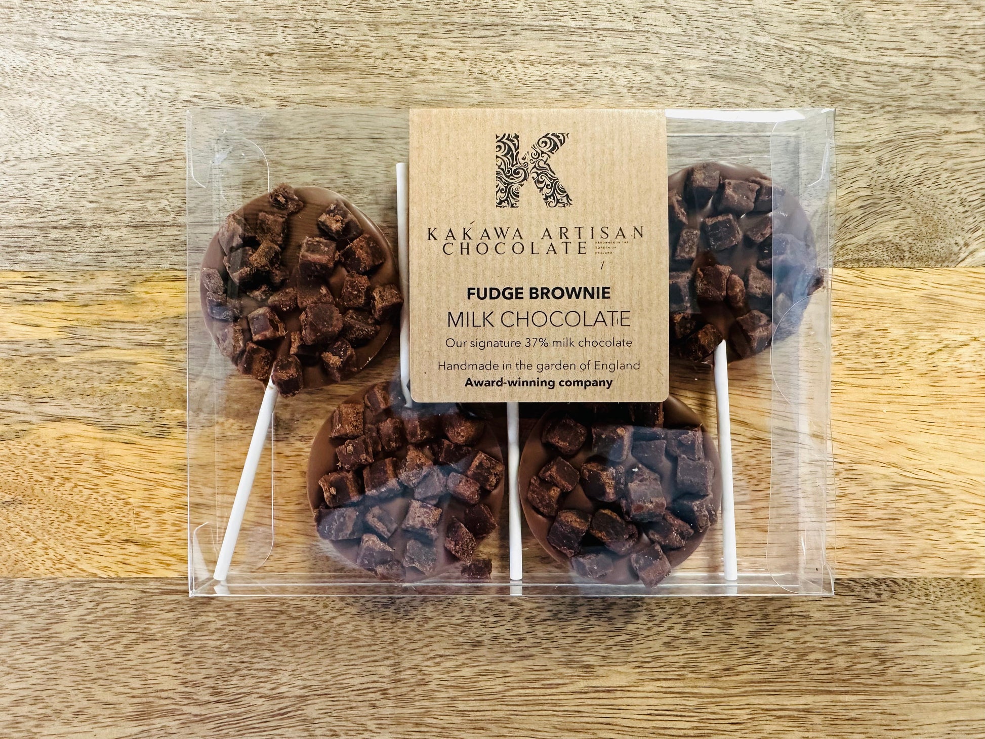 Chocolate fudge brownie lollipops in clear packaging with a Kakawa Artisan Chocolate label on a wooden surface.