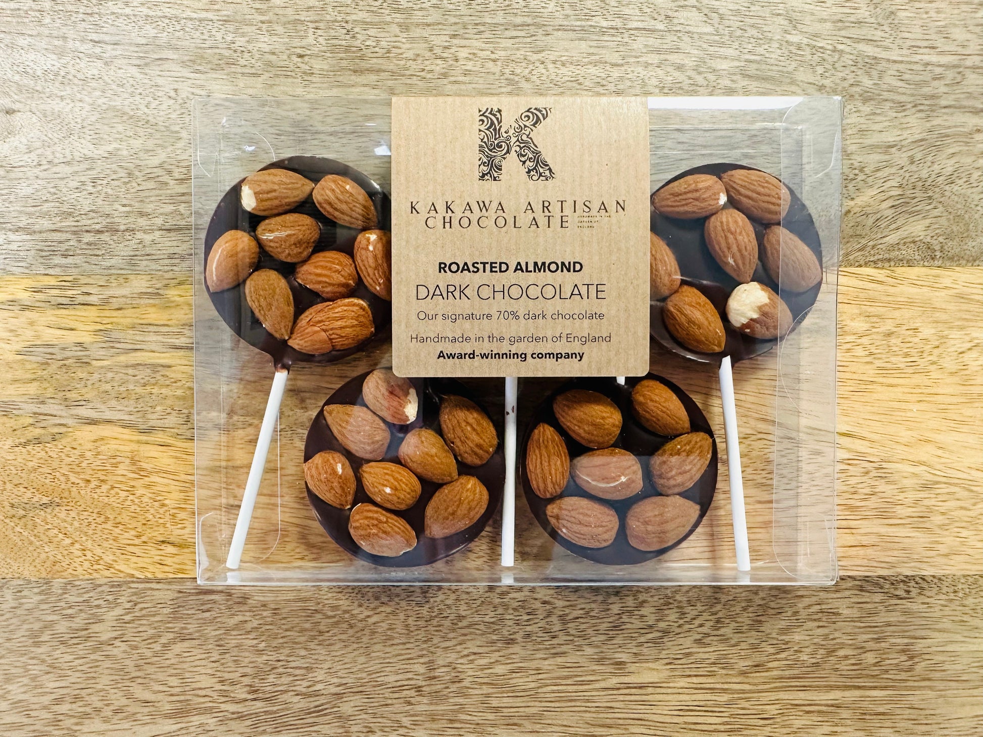 Packaged chocolate lollipops with almonds on a wooden surface, featuring Kakawa Artisan Chocolate branding.